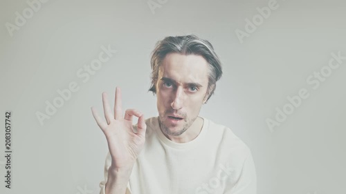 Young man in white long-sleeve shirt making OK hand gesture, approval and consent concept, studio portrait on light gray background, copy space