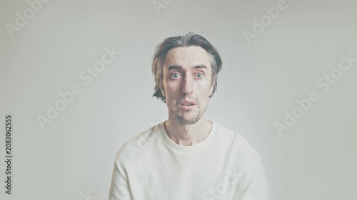 Surprised man looking at camera in studio, sitting against light gray background, wearing plain white long-sleeve shirt, soft even lighting, shallow depth of field