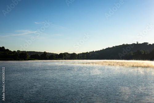 Wide view of a placid lake at daybreak with a band of mist gliding along the distant forested hills and smooth water stretching to the horizon under a pale blue morning sky.