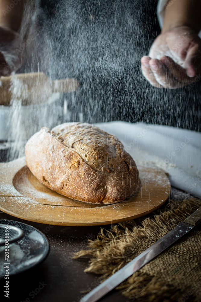 Fototapeta premium Artisan baker hands dusting white flour over hot freshly baked sourdough bread loaf on wooden board. Rustic traditional homemade bakery production concept with dramatic flour splash.
