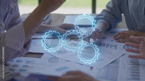 Business people discussing financial charts with digital gears overlay, representing teamwork, data analysis, workflow automation and business process planning.