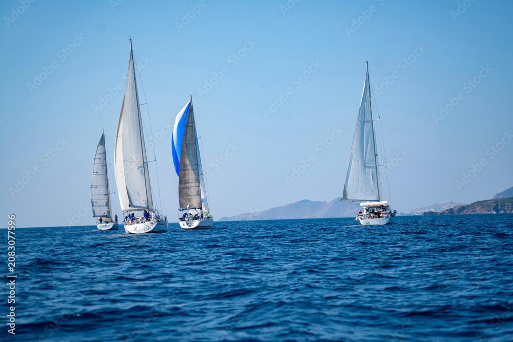 Fototapeta premium Luxury sailing yachts and white boats cruising on the deep blue Mediterranean sea during an active summer regatta ocean vacation with a scenic coastal resort town and mountains in the background