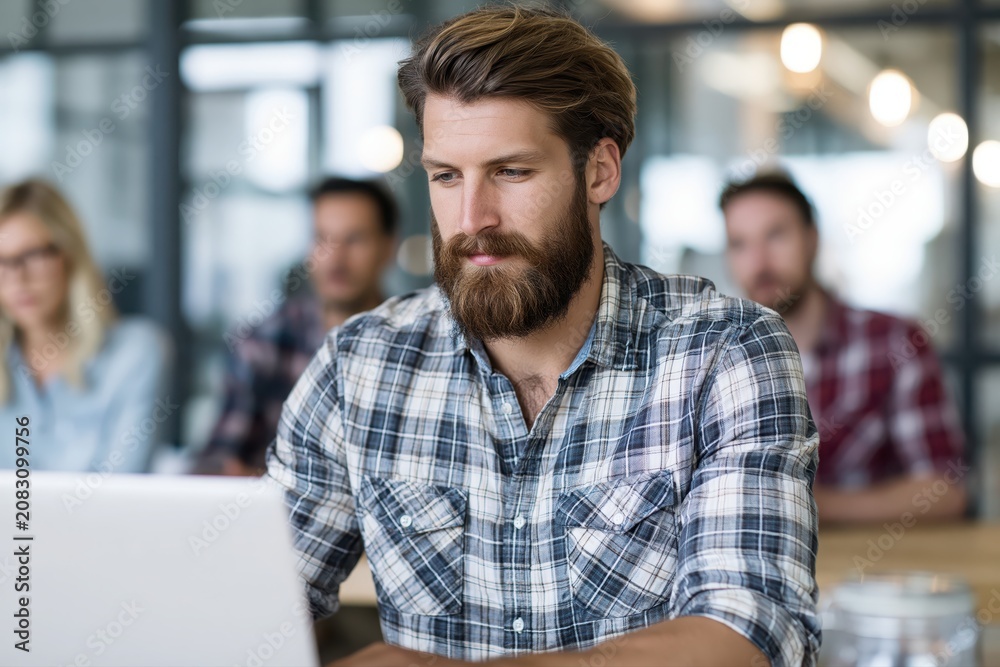 Obraz premium Focused bearded man in plaid shirt working on laptop in modern office with blurred colleagues in background