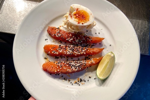 Close-up of smoked salmon slices served with a soft-boiled egg, lime wedge, and sesame seed garnish on a white plate. Fresh seafood dish ideal for brunch, breakfast, healthy eating, gourmet dining, 