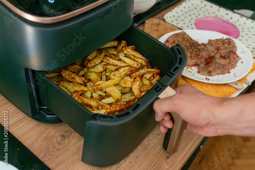 A hand pulls out an air fryer basket filled with golden homemade potato fries seasoned with herbs, with cooked meat on a plate nearby. Useful for home cooking, kitchen appliance, dinner preparation, a
