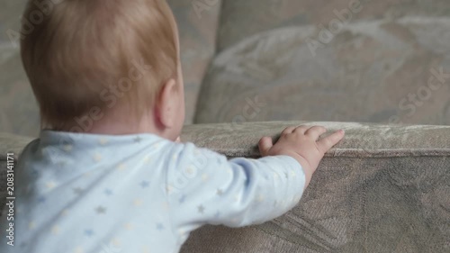 Infant steadies with furniture support. Young child tries to balance while holding onto furniture. Baby in soft clothing works to maintain upright posture using nearby furniture in warm room