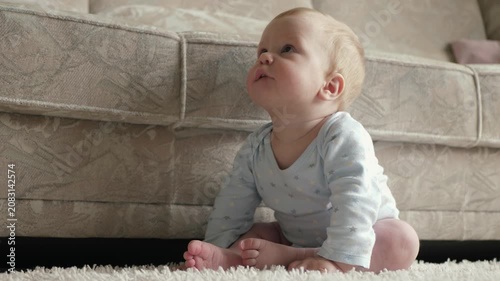 Baby relaxes on rug with curious gaze. Baby sits on plush carpet observing surroundings with gentle curiosity and calmness in warm family setting. Young child in onesie investigates room environment