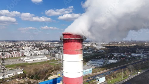 Industrial smokestack emits thick white smoke against a backdrop of urban landscape, showcasing a power plant's operational activity and the surrounding city environment