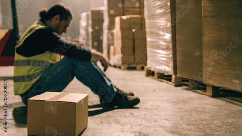A man in a safety vest sits on the floor of a warehouse, leaning on a box, appearing tired or stressed, surrounded by stacked pallets and boxes.