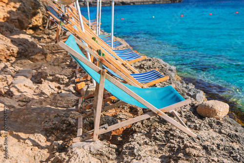 Εκτύπωση καμβά Row of empty deck chairs lines a rocky Mediterranean shoreline beside vivid turquoise water