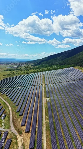 Rows of solar panels spread across a green valley with mountains in the background. The sky shows clouds in a clear day. This site focuses on renewable energy.