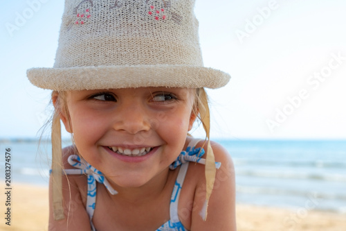 Close up portrait of a smiling toddler girl wearing a white straw sun hat on a beach on a sunny day