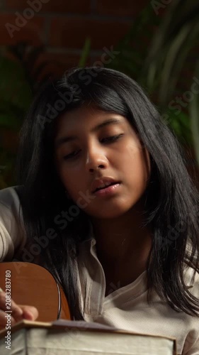 Indian Teenage Girl Playing Guitar While Sitting on a Bean Bag in Cozy Room with Moody Lamp Against Brick Wall, Composing or Writing Music as a Creative Hobby During Free Time at Home