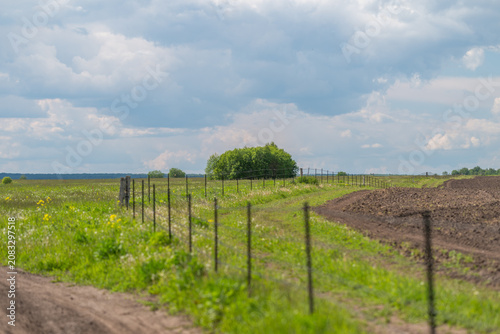 Obraz na plátne Rural dirt road beside fenced green meadow and plowed field under cloudy sky