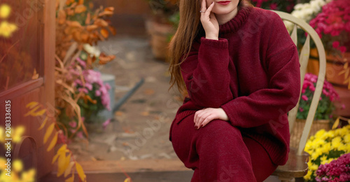 Banner with unrecognizable young woman in a cozy burgundy knitted sweater and trousers sitting on a wooden chair in an autumn garden decorated with colorful chrysanthemums. Copy space on the left