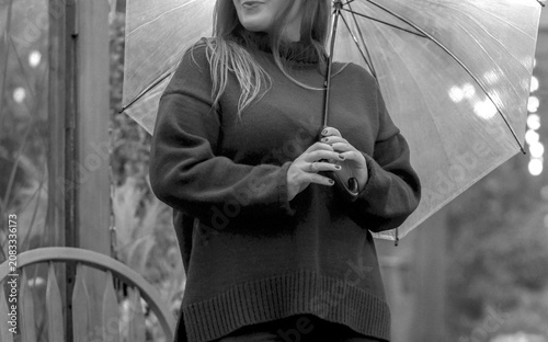 Black and white crop shot of an unrecognizable young woman holding a transparent umbrella in an autumn garden on a rainy day.