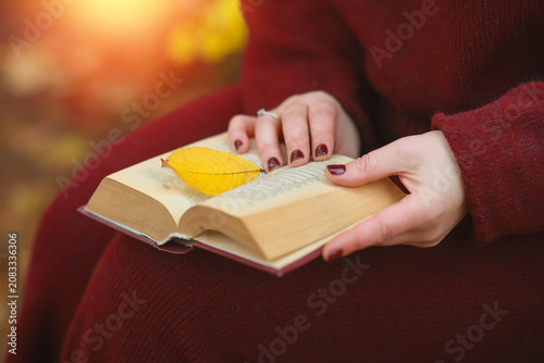 Unrecognizable female reading a book outdoors in a fall park, hands with dark manicure holding pages, warm golden light background.