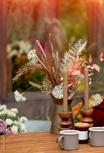 Rustic fall outdoor table setting with warm candles, mugs, an open book, and dried botanical arrangement in a vintage pitcher, autumn lifestyle concept.