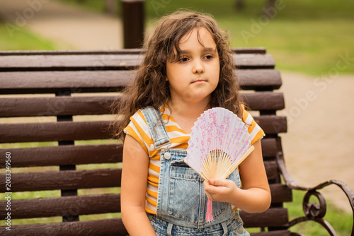 Canvas Print Cute little girl kid child is sitting on a park bench, fanning herself with a decorative fan to cool off