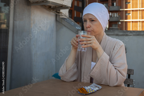cancer patient drinks water after medicine at a sunlit balcony table, with pills in blister packs, representing prescribed therapy, chronic disease care and resilience