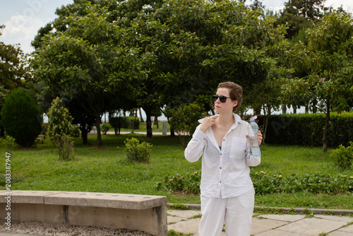 adult cancer survivor pauses with portable water in a public park, symbolizing heat management, energy pacing and daily recovery routines during a warm day of outdoor rehabilitation