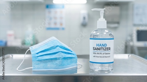 A bottle of hand sanitizer sits next to a blue surgical mask on a white surface. The sanitizer bottle displays text indicating it kills 99.