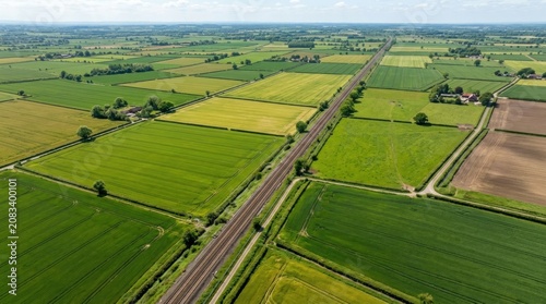 An aerial photograph reveals a vast, undulating landscape of farmland. A long, straight railway line bisects the scene, cutting through fields of vibrant green crops.