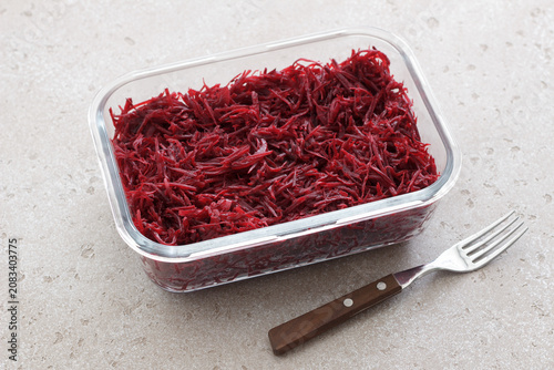 Fresh grated beetroot salad in a glass food container with fork on a light stone surface. Healthy vegetable dish, meal prep ingredient