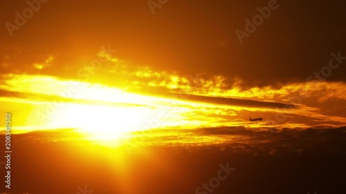 Airplane flying through orange sunset sky with bright sun glowing behind dramatic clouds, silhouette, wide angle real-time shot