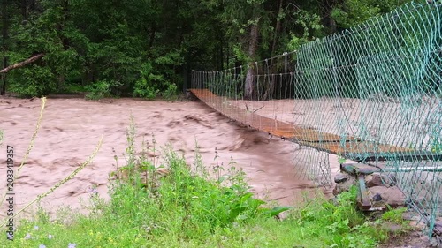 Flash flood and mudflow in Turgen Gorge, Almaty region, Kazakhstan. Raging muddy river current near a suspension bridge. Editorial, travel, and environmental disaster background with copy space.