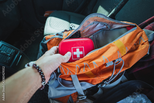 Hand placing red first aid kit into open orange backpack on car seat with hiking gear packed inside, showing travel preparedness, emergency supply storage, and compact organization in natural daylight