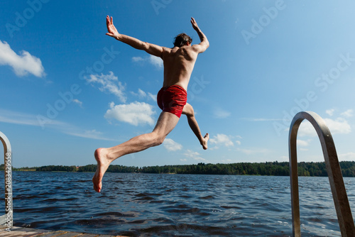 Diver plunges off pier into lake with focused athletic entry on summer day. Shirtless man in red trunks descends midair with concentrated precision outdoors. Powerful plunge creates splash as he
