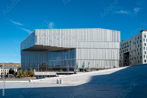 Modern facade of the Oslo Public Library (Deichman Bjørvika) with distinctive vertical white fins and a vast glass wall, overlooking a flag-lined public plaza under a clear sky.