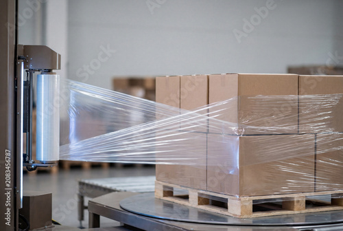 Stretch wrapping machine wraps a pallet stacked with cardboard boxes in plastic film inside a warehouse.
