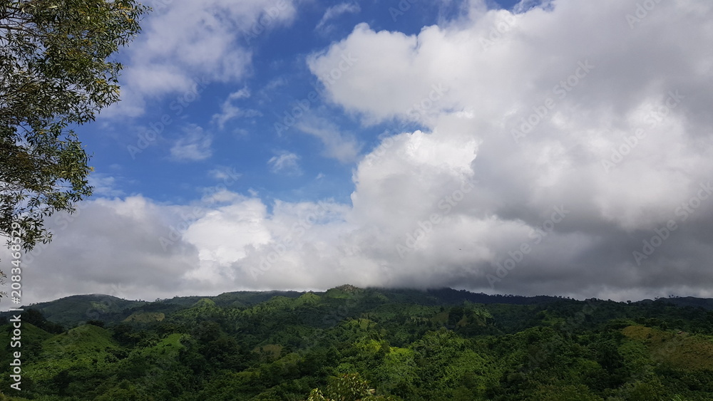 Obraz premium Breathtaking View of Lush Green Tropical Mountains Beneath a Vast Blue Sky with Soft White Clouds on a Sunny Day
