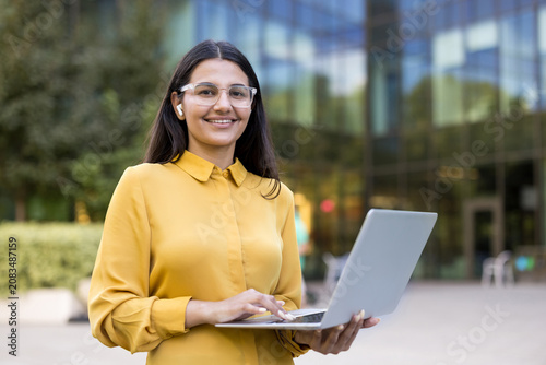 Professional woman wearing glasses and earbuds, smiling while typing on her laptop, conducting remote work or studying online outside a modern office building