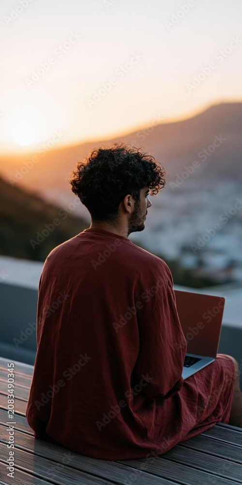 Fototapeta premium Young man sitting with laptop on terrace at sunset overlooking a coastal town for remote work background and travel lifestyle banner
