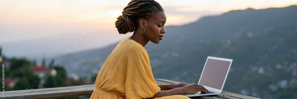 Fototapeta premium Young woman working on laptop outdoors at sunset on balcony with mountain view for remote work banner background