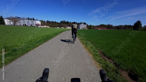 First person pov shot from drop handlebars tracking a road cyclist from behind on an empty asphalt countryside lane surrounded by vibrant green spring fields under a clear blue sky in Bavaria
