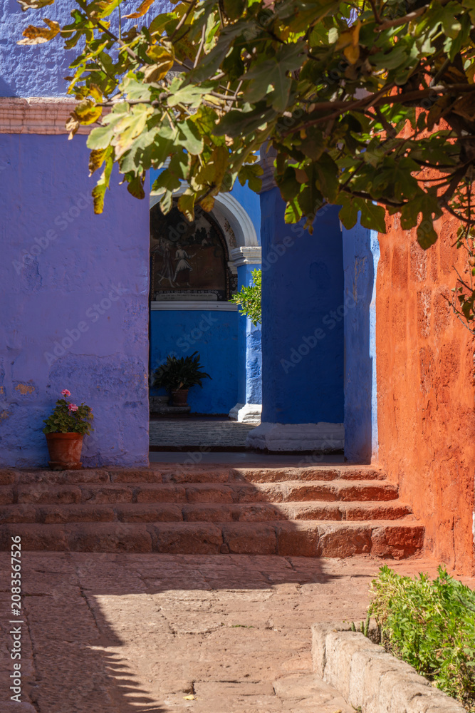 Fototapeta premium A stone pathway and steps lead past contrasting red and vibrant blue sillar stone walls under overhanging tree branches, looking toward a courtyard entrance inside the historic Santa Catalina Monaster