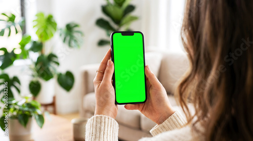 Woman holding mobile phone with green screen mockup in bright cozy living room, person using smartphone for mobile app demo, remote work tasks or social media browsing at home.
