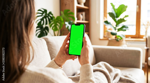 Woman holding mobile phone with green screen mockup while relaxing on couch at home, using smartphone for leisure, social media browsing or mobile app demo in cozy living room.
