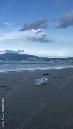 Discarded clear plastic water bottle resting on wet sandy shoreline beneath wide blue sky with distant island, low surf, foam, reflective tide marks, and quiet coastal pollution scene