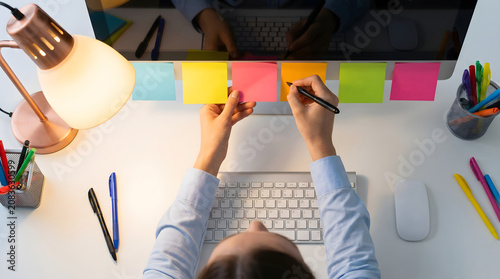 Woman writing notes and attaching colorful sticky notes on office desk with keyboard and lamp