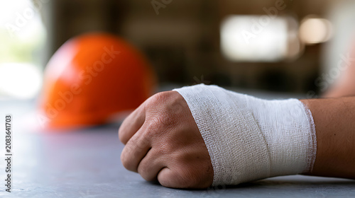 Injured hand with a bandage and an out-of-focus hard hat, symbolizing a workplace accident or an unfortunate incident in a work environment