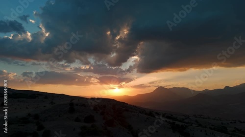 Dramatic sky with dark clouds. Sun low on horizon. Distant mountains outline the view. Desolate terrain stretches toward the setting sun. Serene atmosphere captures natural beauty