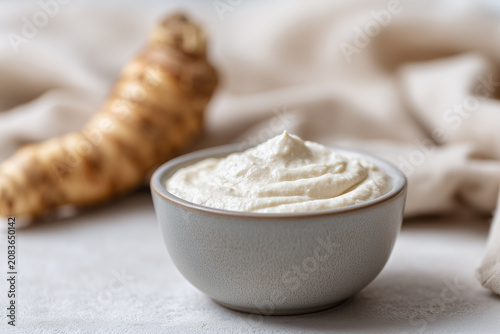Fresh grated horseradish sauce in a ceramic bowl with raw root on bright background