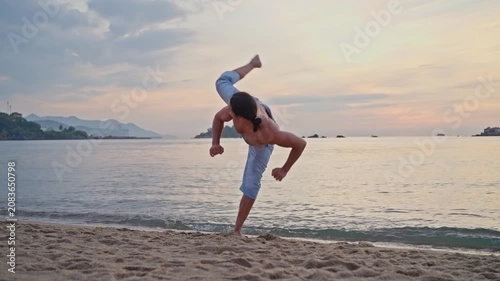 Shirtless man practicing controlled martial arts and fitness movements on sandy beach at sunset with rolled pants. Slow motion roundhouse kick