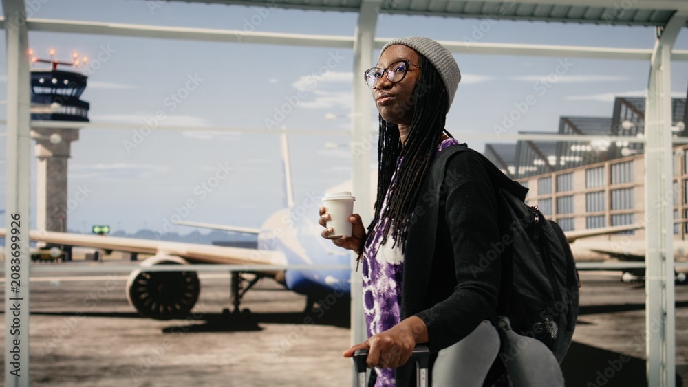 Obraz premium Woman in busy airport terminal traveling to destination with coffee cup in hand, Young african american girl carrying trolley in airport hallway, holding drink container for travel concepts.