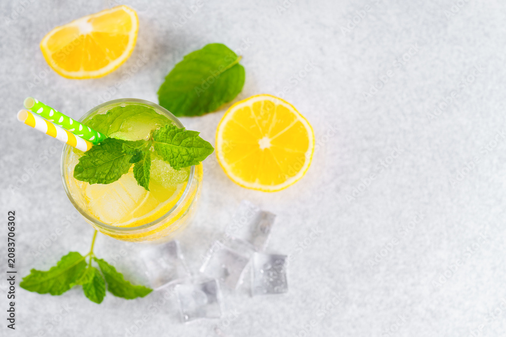 Fototapeta premium Top view of refreshing cold icy beverage, homemade lemonade with fresh lemon slices, mint leaves, ice cubes wtih soda water and condensation in glass on gray background with copy space for text.